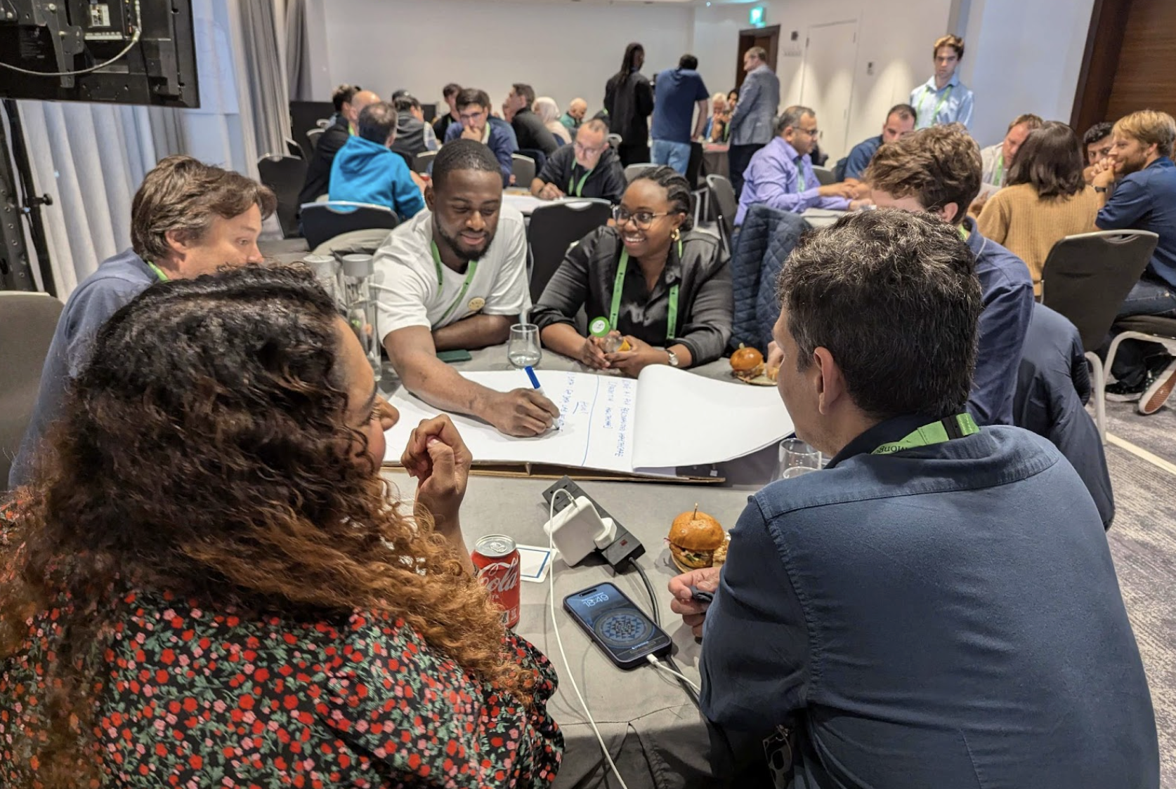 Photo of .local London AI Build Together attendees brainstorming AI solutions for the healthcare industry. The group is sitting around a table and writing down ideas on a giant notepad. The room is filled with more tables of other groups doing the same thing.